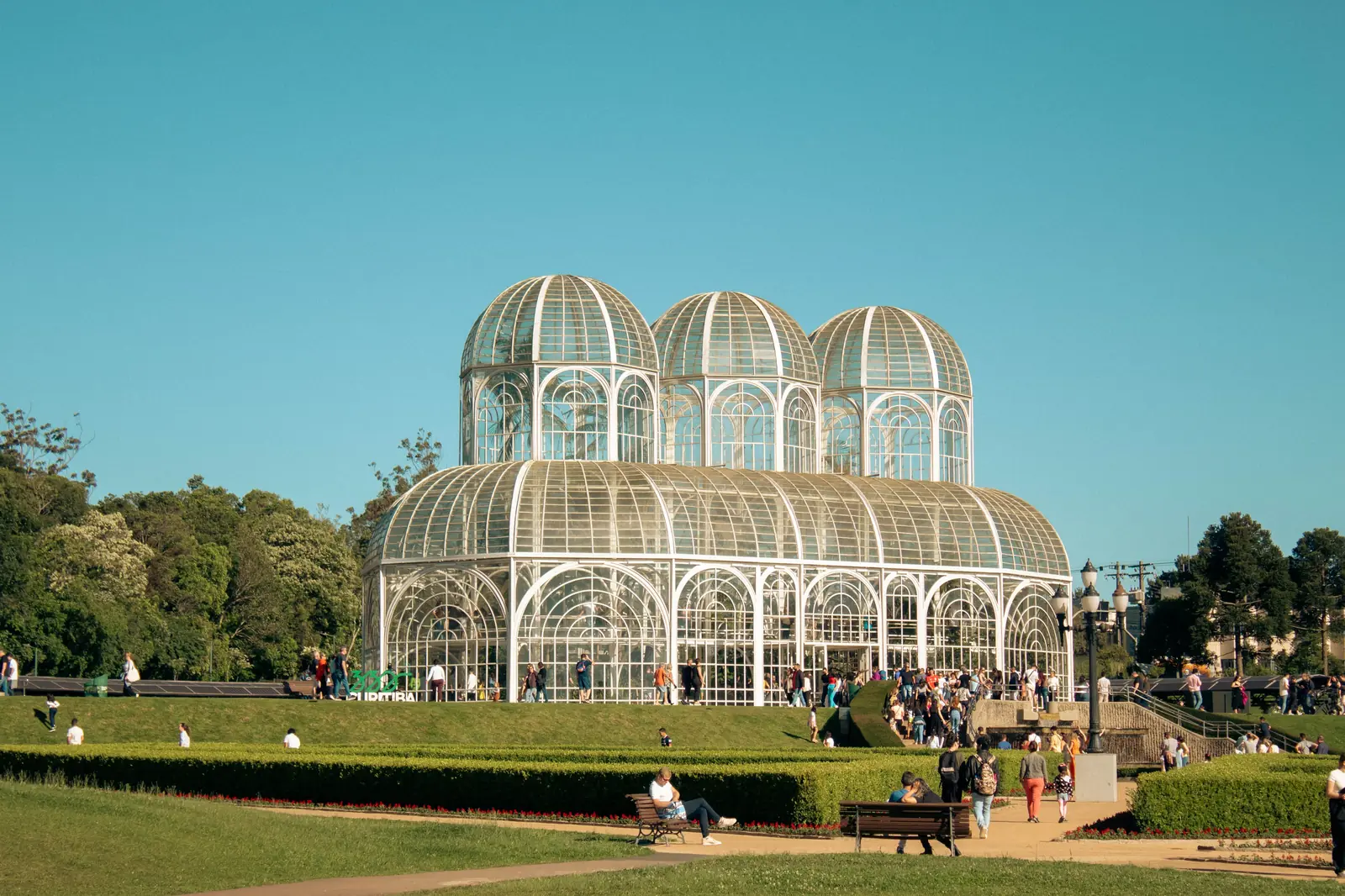 Estufa do Jardim Botânico de Curitiba, com estrutura de vidro e ferro branco e o jardim geométrico em primeiro plano
