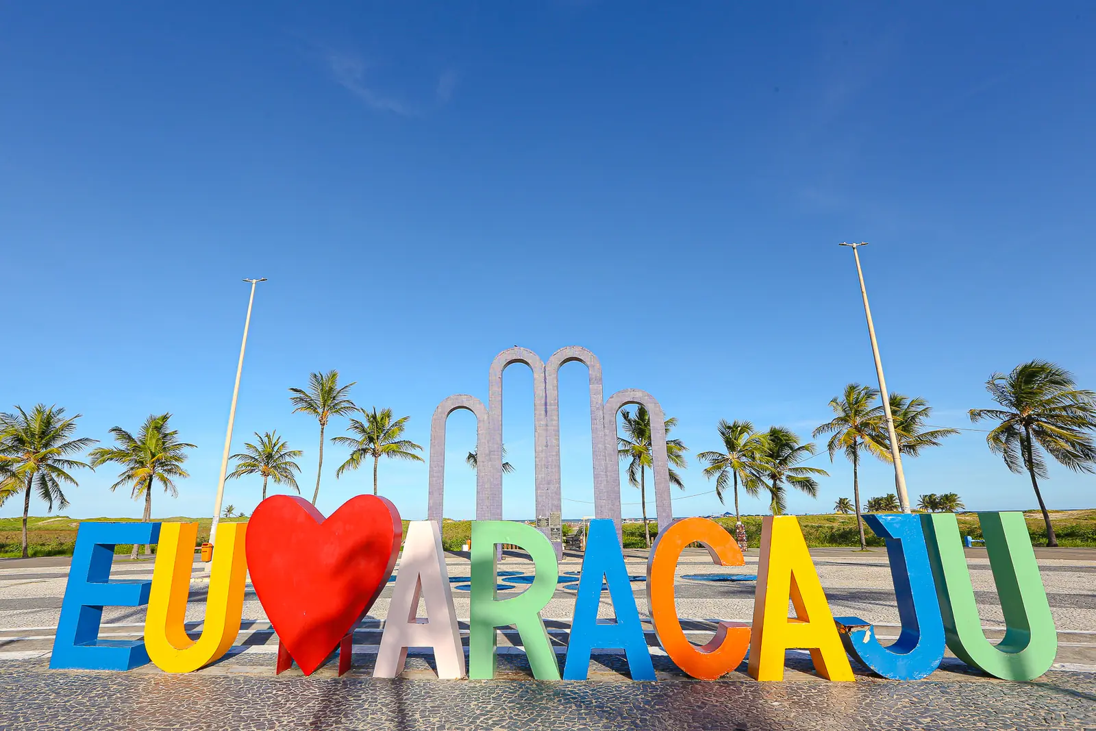 Letreiro "Eu amo Aracaju" na orla de Atalaia, com os coqueiros e os arcos do Passeio do Caranguejo ao fundo