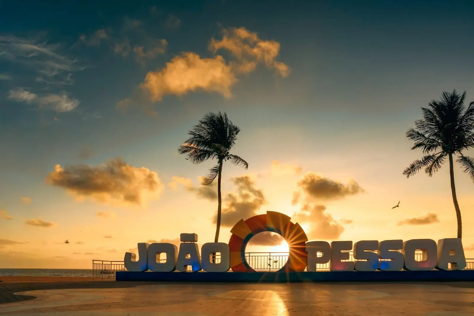 Letreiro de João Pessoa na orla ao pôr do sol, entre dois coqueiros, com o sol atravessando o círculo central