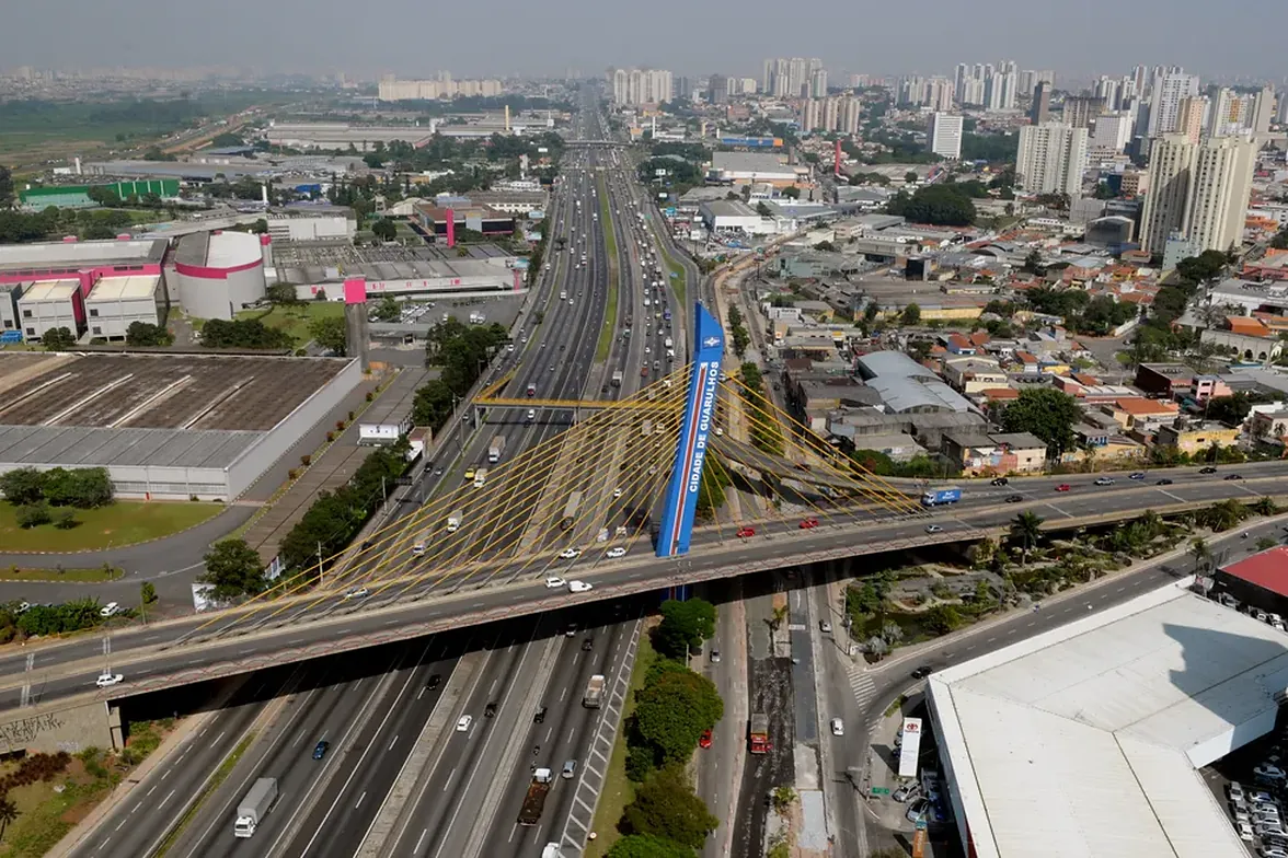 Ponte estaiada Cidade de Guarulhos, com o mastro azul e os cabos amarelos, cruzando a rodovia e o centro da cidade ao fundo